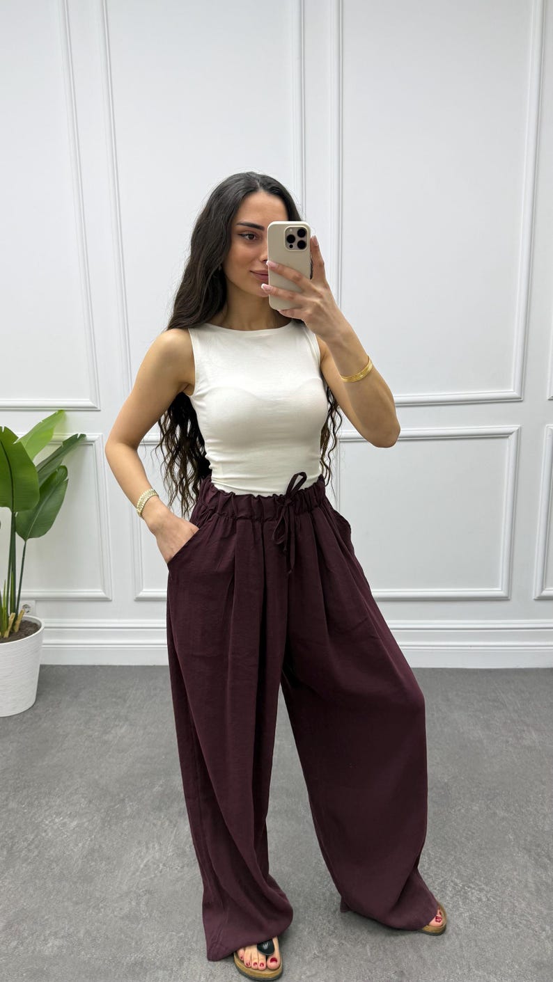 Op de afbeelding: Een vrouw draagt een witte mouwloze top en een wijde, bordeauxrode broek. De broek heeft een taille met trekkoord en zakken. Ze houdt een telefoon vast en draagt gouden armbanden.
