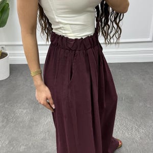 Op de afbeelding: Een mouwloze witte top gecombineerd met een hoog getailleerde, bordeauxrode broek met wijde pijpen. De broek heeft zakken en een geplooid tailleband. De outfit wordt gecompleteerd met sandalen. Gouden armbanden sieren de polsen.