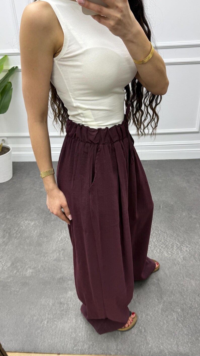 Op de afbeelding: Een mouwloze witte top gecombineerd met een hoog getailleerde, bordeauxrode broek met wijde pijpen. De broek heeft zakken en een geplooid tailleband. De outfit wordt gecompleteerd met sandalen. Gouden armbanden sieren de polsen.