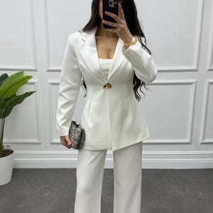 May include: A white pantsuit featuring a tailored blazer with a single gold button closure and wide-leg pants. The outfit includes a matching top and is paired with a small, patterned clutch. The ensemble is a modern take on classic tailoring.