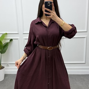 May include: A long, button-down burgundy dress with a collared neckline and long sleeves. The dress is cinched at the waist with a brown belt. The model is holding a phone in front of their face. The dress is made of a flowing fabric.