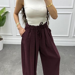 Op de afbeelding: Een damesoutfit met een witte mouwloze top en een bordeauxrode broek met wijde pijpen en een taille met trekkoord. De outfit wordt gecombineerd met sandalen en gouden armbanden. De broek heeft zakken en een relaxte pasvorm, geschikt voor casual wear.