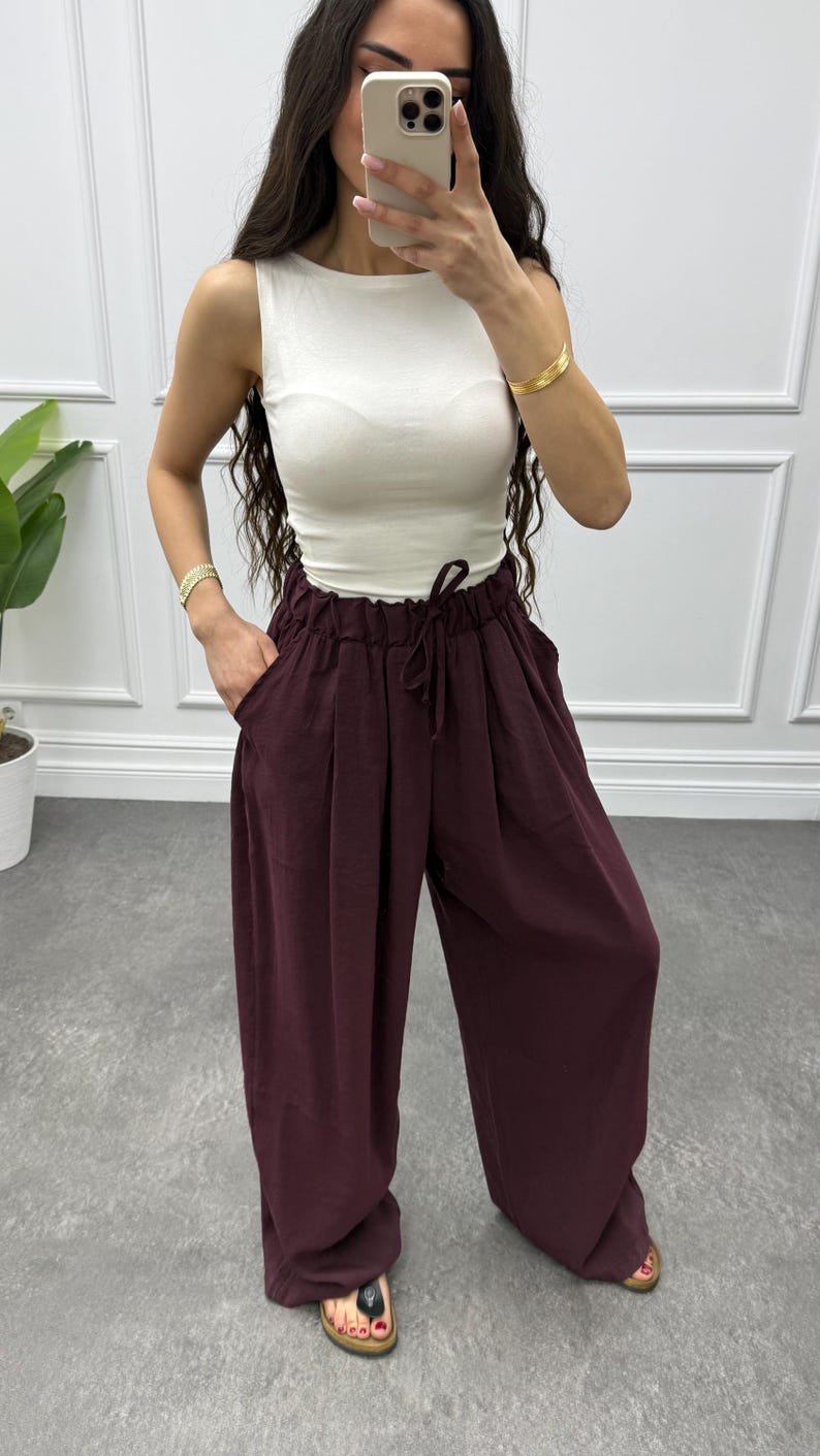 Op de afbeelding: Een damesoutfit met een witte mouwloze top en een bordeauxrode broek met wijde pijpen en een taille met trekkoord. De outfit wordt gecombineerd met sandalen en gouden armbanden. De broek heeft zakken en een relaxte pasvorm, geschikt voor casual wear.