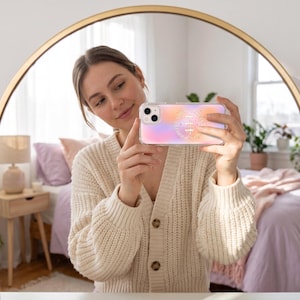 Könnte beinhalten: Eine transparente Smartphone-Hülle mit einem Farbverlauf von zartem Rosa zu Orange. In der Mitte der Hülle befindet sich eine kreisförmige goldfarbene Grafik mit der Zahl 411, umgeben von kleinen Sternsymbolen und kreisförmigem Text.