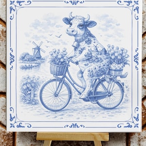 Op de afbeelding: Blauw-witte keramische tegel met een koe op een fiets, met manden vol tulpen. Het lichaam van de koe is versierd met een molen en een landschap. De tegel heeft een decoratieve rand en wordt tentoongesteld op een klein houten ezel.
