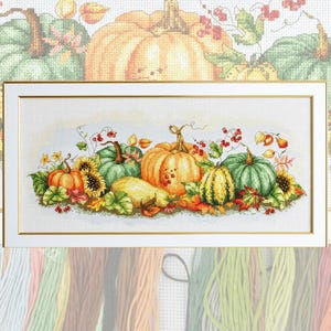 May include: A cross-stitch embroidery pattern featuring a horizontal arrangement of orange, yellow, and green pumpkins, gourds, sunflowers, and red berries. The design is set against a white fabric background and framed in a thin gold-toned border.