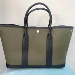 Pode incluir: Uma bolsa tote de lona verde-oliva com acabamento em couro preto, duas alças superiores e um fecho de botão de pressão de metal prateado no centro. A bolsa possui uma base estruturada e ilhós de metal nas laterais.