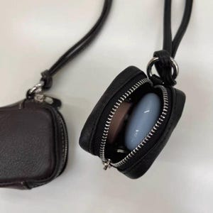May include: Two small black square pouches with silver zippers and long black neck straps. One pouch is closed, while the other is open, revealing a smooth light blue oval object and a flat pink circular object stored inside the main compartment.