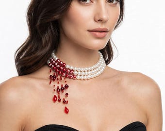 May include: A multi-strand choker necklace featuring white pearls that transition into deep red pearls on one side. A cluster of dangling red beads and teardrop-shaped charms hangs from the side of the necklace, creating a layered effect.