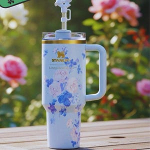 May include: A light blue insulated travel mug with a handle, decorated with a floral design in pink and blue. It includes a straw and a decorative top. The mug has the brand name "STANLEY" and the text "LOVESHACKE".
