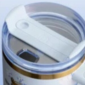 May include: Close-up of a white and clear travel mug lid. The lid has a sliding mechanism to open and close the drinking spout. The mug's body is visible, featuring a gold band and floral design.