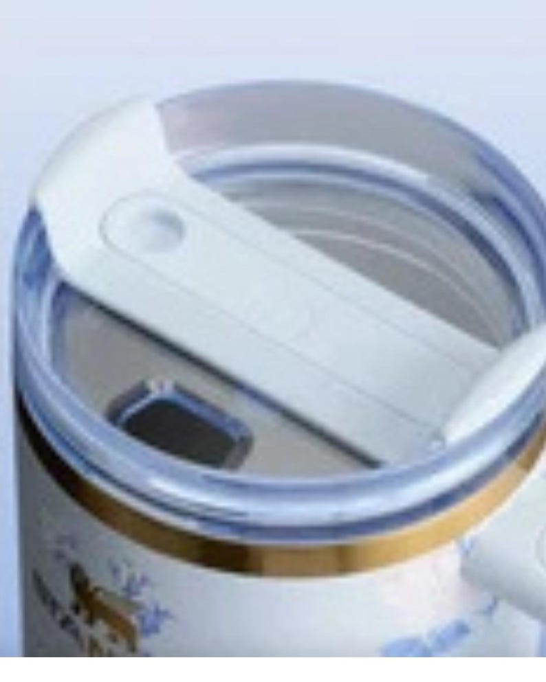 May include: Close-up of a white and clear travel mug lid. The lid has a sliding mechanism to open and close the drinking spout. The mug's body is visible, featuring a gold band and floral design.