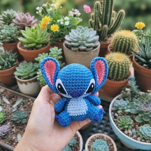 May include: A blue crochet Stitch doll with pink inner ears and black eyes is held in front of a collection of potted succulents and cacti. The doll has a light blue belly and is made of yarn.
