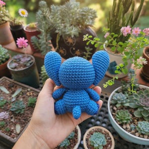 May include: A handmade, blue crochet Stitch doll with large ears, arms, and legs. The plush toy is held in front of a background of potted plants and flowers, including succulents and daisies. The doll is approximately 5 inches tall.