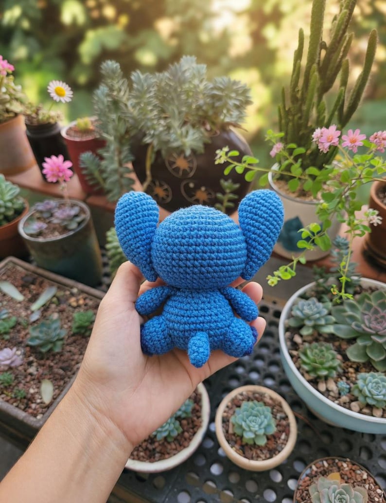 May include: A handmade, blue crochet Stitch doll with large ears, arms, and legs. The plush toy is held in front of a background of potted plants and flowers, including succulents and daisies. The doll is approximately 5 inches tall.