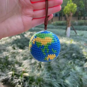 Pode incluir: Enfeite de globo terrestre de crochê, com oceanos azuis, continentes verdes e amarelos e uma alça de couro marrom para pendurar. A esfera feita à mão está suspensa em um fundo verde desfocado.