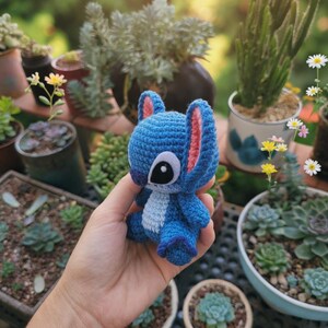 May include: A handmade blue crochet Stitch doll with pink inner ears and a white chest. The doll is held in a hand, with a blurred background of potted plants and succulents.