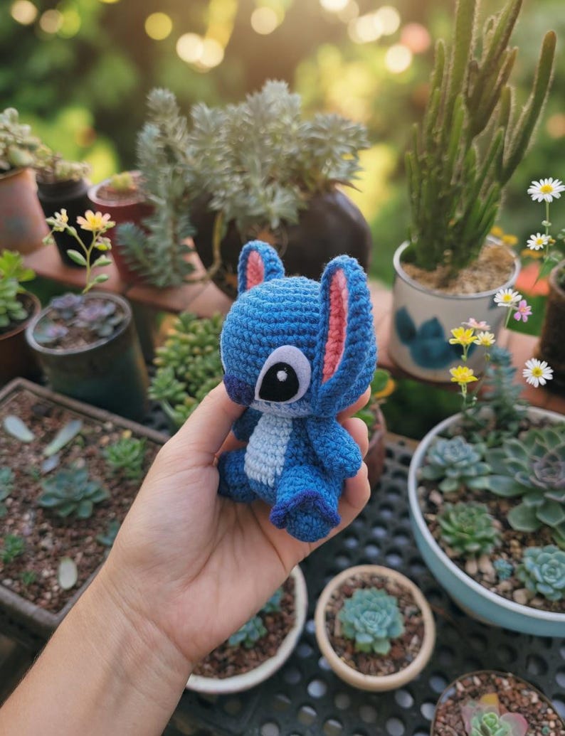 May include: A handmade blue crochet Stitch doll with pink inner ears and a white chest. The doll is held in a hand, with a blurred background of potted plants and succulents.