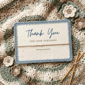 May include: A cream-colored card with blue stitched edges and the words "Thank You" in elegant script. The card is tied with twine and rests on a crocheted blanket with flower accents. The text "FOR YOUR PURCHASE" and "We hope you love it!" are also visible.