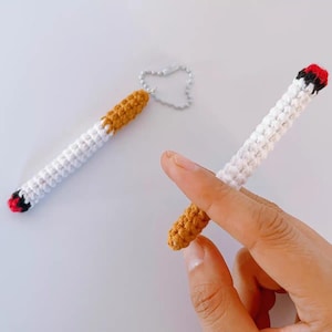 May include: Crocheted cigarette keychain with a white body, brown filter, and red and black tip. One keychain is attached to a silver chain. The other is held by a hand. The items are on a white background.