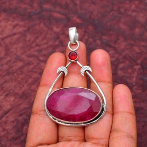 Può includere: Un ciondolo in argento con una grande pietra preziosa di colore rosa scuro, di forma ovale e sfaccettata, incastonata in una montatura di metallo. Sopra la pietra principale, una pietra rossa più piccola, rotonda e sfaccettata, con due dettagli in argento a forma di mezzaluna.