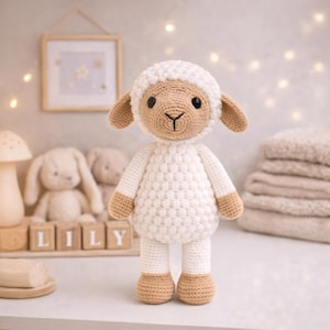 May include: A handcrafted crochet lamb toy, featuring a white body and head, with tan face, ears, and feet. The doll has a textured stitch pattern and is standing. The background includes a framed picture, wooden blocks spelling "LILY", and other soft toys.