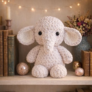 Könnte beinhalten: Ein handgefertigtes, hellbeige gehäkeltes Elefanten-Plüschtier mit großen Ohren, einem Rüssel und schwarzen Knopfaugen. Der Elefant sitzt auf einem Holzregal mit Büchern und dekorativen Gegenständen. Im Hintergrund befinden sich eine Blumenvase und Lichterketten.