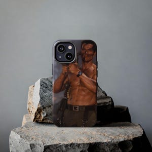May include: A smartphone case featuring a photographic print of a shirtless, muscular individual with dark face paint, wearing a black belt with ammunition and a wristwatch, holding a large firearm in a jungle-themed action movie scene.