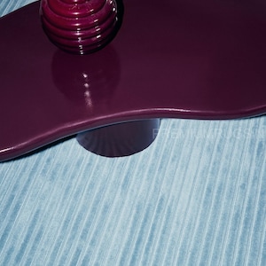 May include: A modern, dark purple coffee table with a unique, wavy shape. The table has a single, cylindrical leg and a glossy finish. A small, round, dark purple vase with dried flowers sits on top. The table rests on a light blue, ribbed rug.