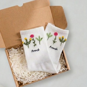 Puede incluir: Un par de calcetines blancos con diseños florales bordados en una caja de cartón llena de papel de relleno. Los calcetines presentan flores rosas, amarillas y verdes, con la palabra "Ammoh" bordada debajo.