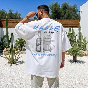 Könnte beinhalten: Ein weißes Oversize-T-Shirt mit einem Grafikdruck auf der Rückseite. Das Design zeigt eine Bierflasche und einen Bierkrug mit dem Text MALLE die Lage ist... ESKALATIONSBEREIT in blauer und schwarzer Tinte für einen lässigen Look.
