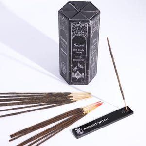 Op de afbeelding: Een zwarte zeshoekige doos met wierookstokjes met de woorden "Success" en "Seek Smudge Incense" erop gedrukt. Er liggen verschillende wierookstokjes, sommige met gouden punten en rode uiteinden, samen met een zwarte wierookhouder met de tekst "ANCIENT WITCH".