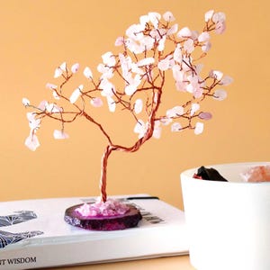 Peut inclure: Une sculpture décorative en forme d'arbre avec un tronc et des branches en fil de cuivre, ornée de feuilles de pierres précieuses roses et blanches. L'arbre repose sur une base violet foncé et rose, sur un livre blanc. Un récipient en céramique blanche est au premier plan.