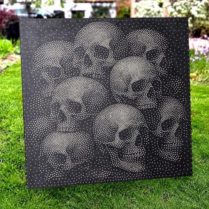 Op de afbeelding: Een vierkant zwart paneel met een pointillistisch ontwerp bestaande uit talloze kleine witte stippen. De rangschikking van deze stippen vormt de duidelijke contouren van meerdere menselijke schedels die over het hele paneel zijn gegroepeerd.