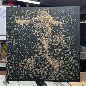 Op de afbeelding: Een vierkant zwart wandkunstwerk met een pointillistisch portret van een stier, gemaakt met talloze kleine goudkleurige stippen. De dichtheid van de stippen varieert om de vorm, de hoorns en de gelaatstrekken van de stier te vormen.