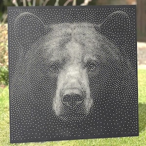 Op de afbeelding: Een vierkant zwart paneel met een pointillistische illustratie van een berenkop, gemaakt met talloze kleine witte stippen. De dichtheid van de stippen varieert om de gedetailleerde kenmerken van de beer te vormen, inclusief de ogen, neus en oren.