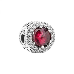 May include: A round silver charm bead featuring a large, faceted, deep red center stone surrounded by a halo of small, clear, sparkling stones. The side of the silver bead is engraved with the text PANDORA and S925 ALE.