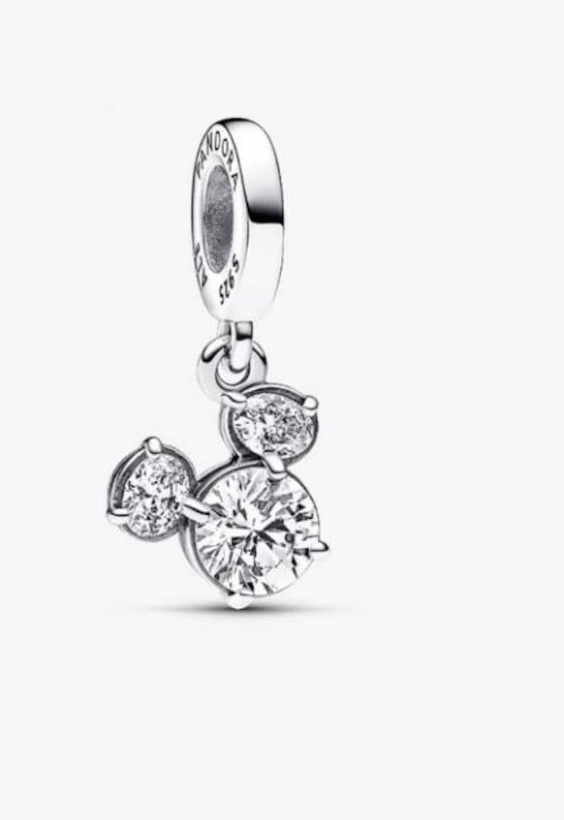 May include: A silver dangle charm featuring three round, clear, faceted stones arranged in a cluster resembling a mouse head silhouette. The top bail is engraved with the text PANDORA and S 925, indicating the metal composition and brand.
