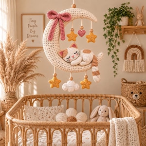 Peut inclure: Un mobile en crochet composé d'un grand croissant de lune crème avec un nœud rose, un chat calico endormi, ainsi que des étoiles jaunes, un cœur rose et un nuage blanc suspendus. Le mobile est fixé par une cordelette avec des perles en bois.