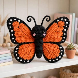 May include: A crocheted monarch butterfly plush toy featuring a black body with pink cheeks and large black eyes. The wings are vibrant orange with black borders and white spots. Two black curled antennae extend from the top of the head.