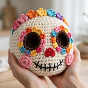 May include: A cream-colored crocheted skull featuring dark brown eye sockets surrounded by multi-colored floral petals in pink, yellow, orange, and blue. It has a red heart-shaped nose and a black stitched mouth with pink circular cheek accents.