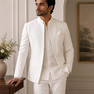 Könnte beinhalten: Ein weißer Blazer mit Stehkragen, getragen über einem passenden weißen Hemd mit Knopfleiste und einer weißen Hose. Der Blazer hat eine klare, strukturierte Silhouette mit einer Brusttasche, in der ein weißes Einstecktuch steckt.