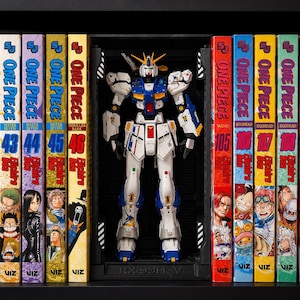 May include: A white, blue, and yellow RX-93ff-v Gundam model figure standing in a black display frame, positioned between several One Piece manga volumes on a shelf. The figure features a detailed mechanical design with a V-fin head crest.