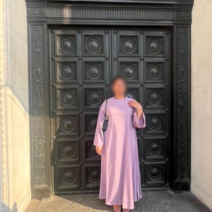 May include: A long, flowing lavender dress with wide sleeves. The dress is worn with a small black purse. The background features a large, ornate black door with decorative panels and a stone facade.