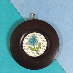 May include: A small, round, wooden ornament with a dark brown finish. The ornament has a clear glass circle in the center with a pressed blue flower inside. The glass circle is surrounded by a printed page with text that reads "in over the beited to his ," Ja with wor him; "a suppose earing whi ent teas? He looke pid!" she pecan-a scarlet-cheeke ing dow"