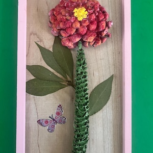 May include: A framed floral art piece featuring a large, textured red and yellow flower, green leaves, and a butterfly. The flower is the focal point, set against a light wood background within a pink frame. The butterfly has purple and red details.