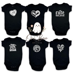 May include: Six black baby onesies featuring white graphic designs. The designs include phrases such as "Dead Cute," "Cute but Feral," and "Too Cute to Spook," alongside heart and ghost illustrations. The brand name "Ori Boo Studio" is also visible.