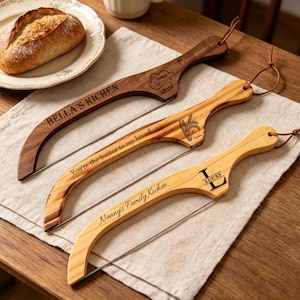 May include: Three wooden bread slicers with stainless steel blades are displayed on a linen cloth. The slicers are engraved with text, including "BELLA'S KICHEN", "You're the butter to my bread", and "Nanny's Family Kichen". Each has a leather loop for hanging.