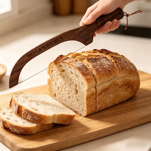 Peut inclure: Un coupe-pain en bois avec l'inscription "BELLA'S KICHEN" coupe une miche de pain sur une planche en bois. Le pain est tranché. Le coupe-pain a une forme incurvée et une lame en fil.