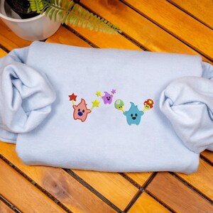 May include: A light blue sweatshirt with embroidered cartoon characters. The design features a pink character holding a red star, a purple character, a green character, and a blue character holding a mushroom.